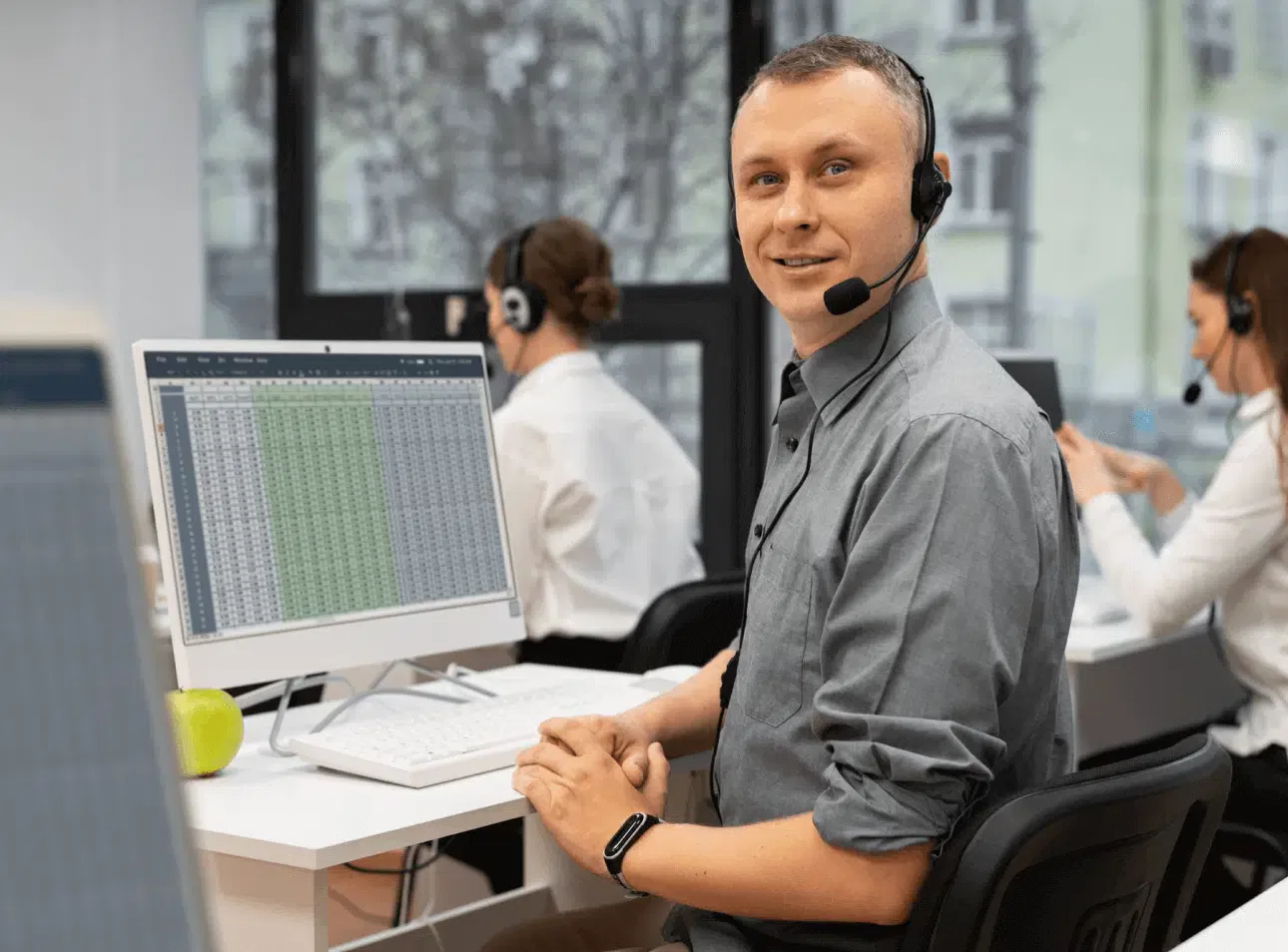 A man wearing a headset smiles at a desk with a computer displaying a spreadsheet, likely analyzing data for managed web hosting solutions. Two people work at computers in the background, contributing to the efficient team effort.