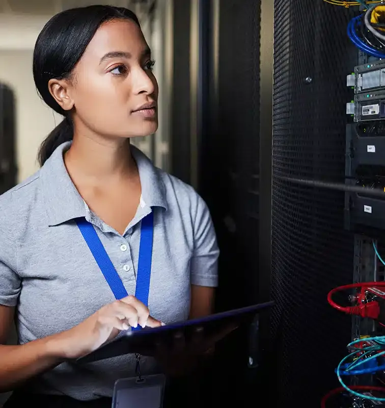 A woman in a server room checks equipment related to managed web hosting while holding a tablet. She is wearing a gray polo shirt and a blue lanyard.